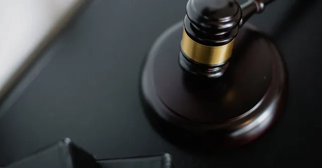 A wooden gavel with a gold band rests atop a matching block, placed on a dark surface.