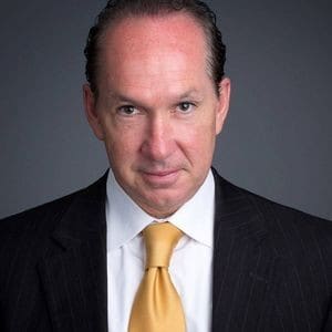 A man in a suit and yellow tie looks directly at the camera against a gray background.