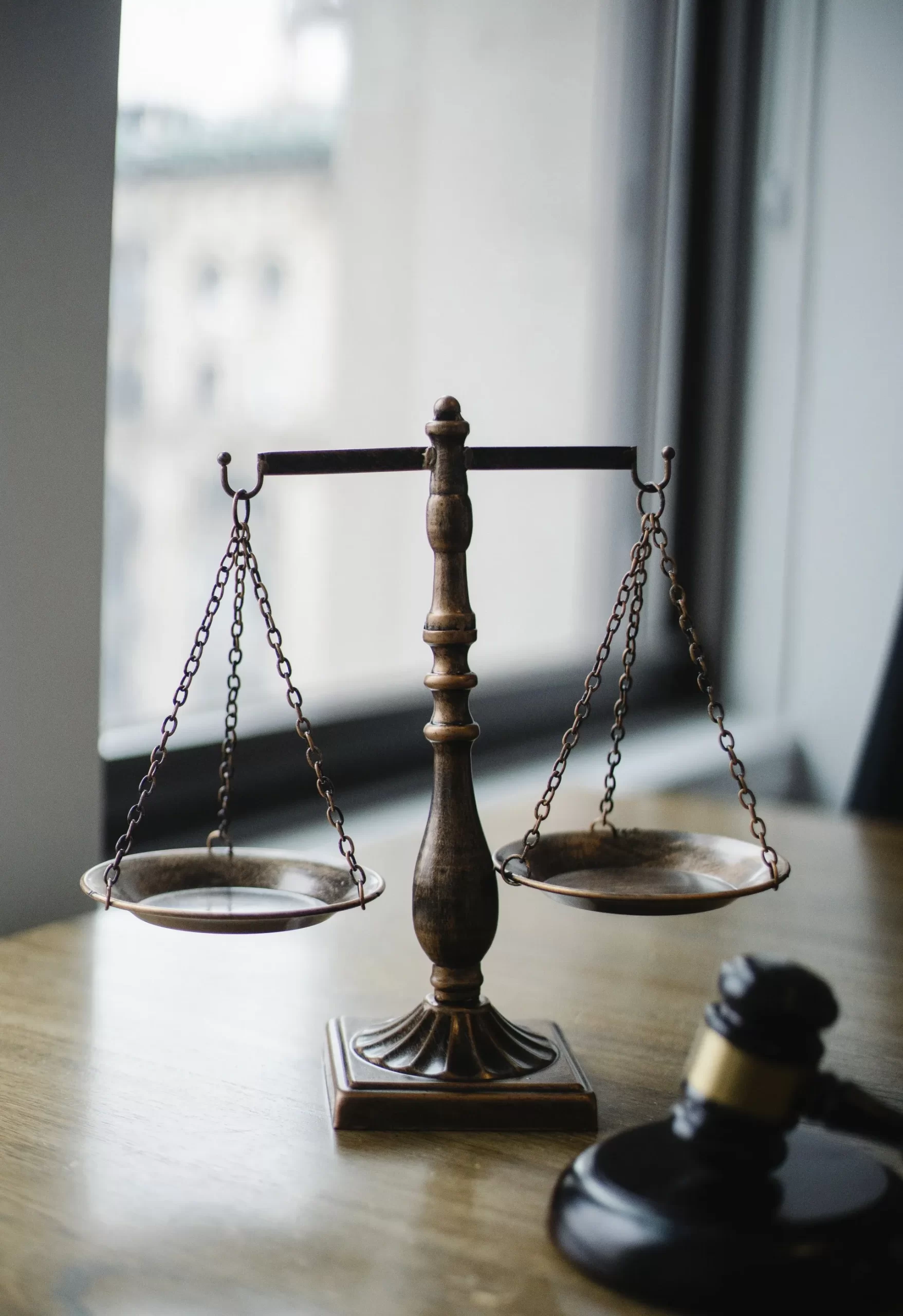 Bronze balance scales on a wooden desk near a window, with a judge's gavel beside them.