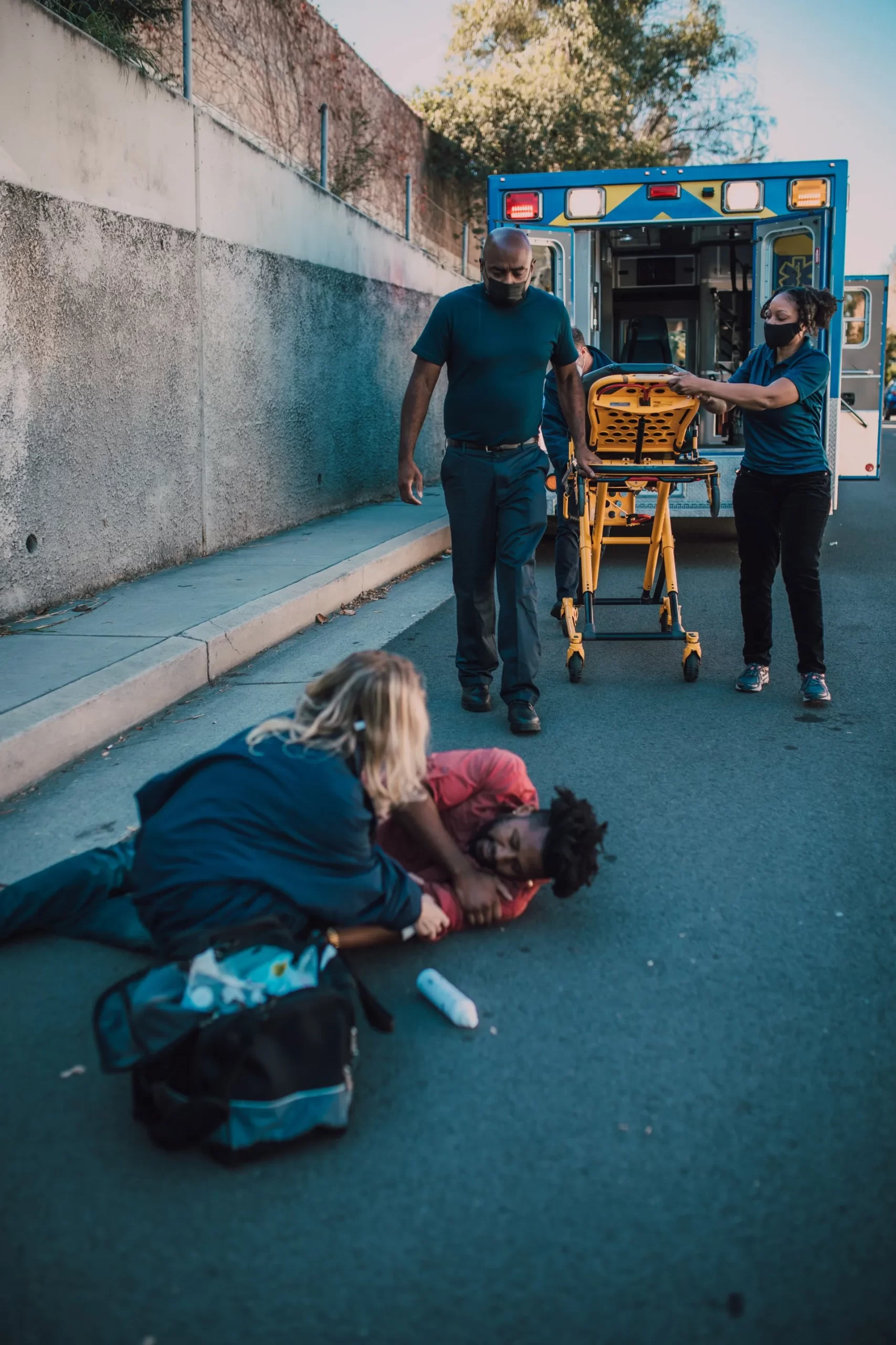 Paramedics assist a person lying on the ground beside an ambulance. A stretcher is being prepared.