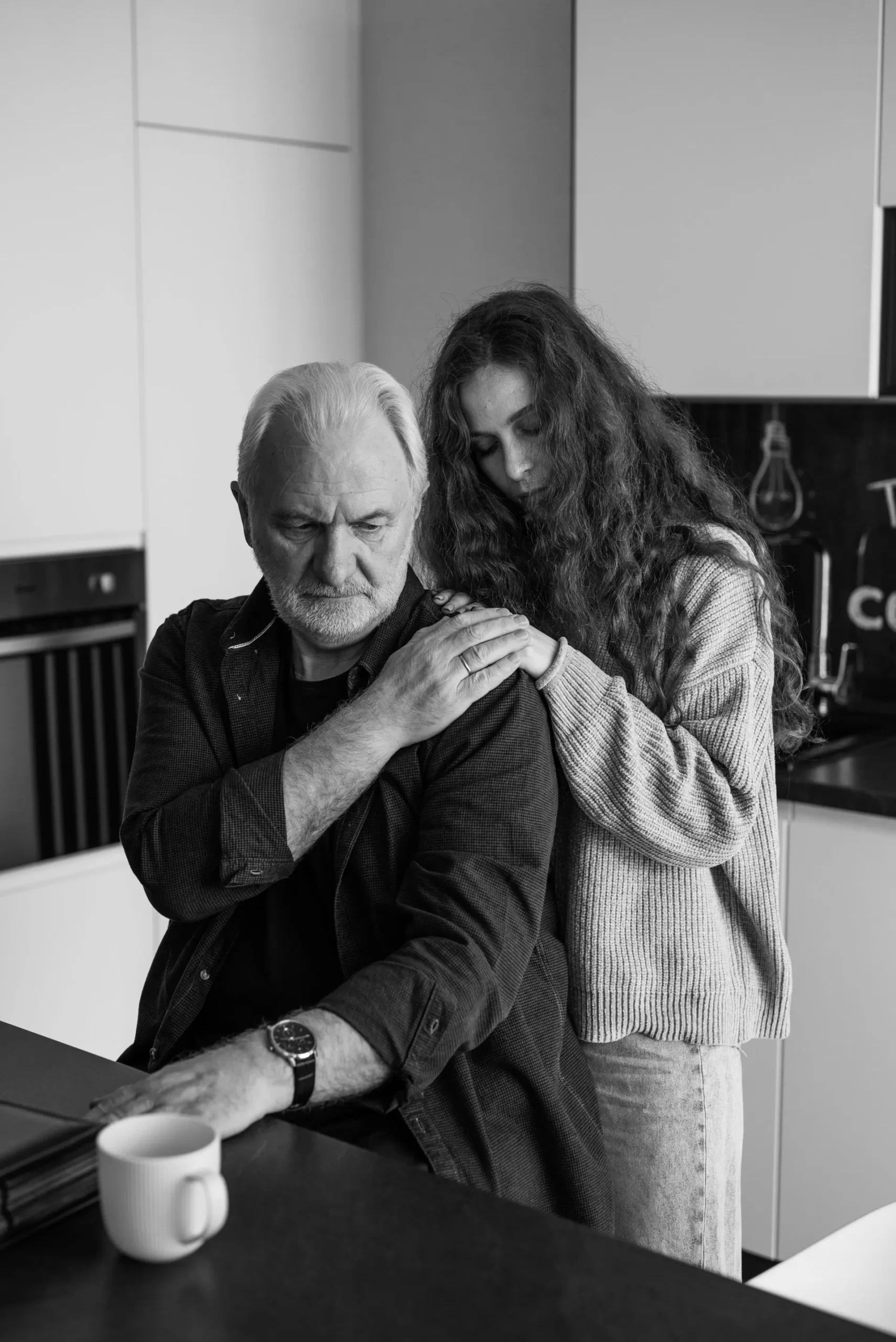 An elderly man sits at a kitchen counter with a mug, while a young woman stands behind him, gently touching his shoulder. They appear thoughtful in a modern kitchen setting. Black and white photo.