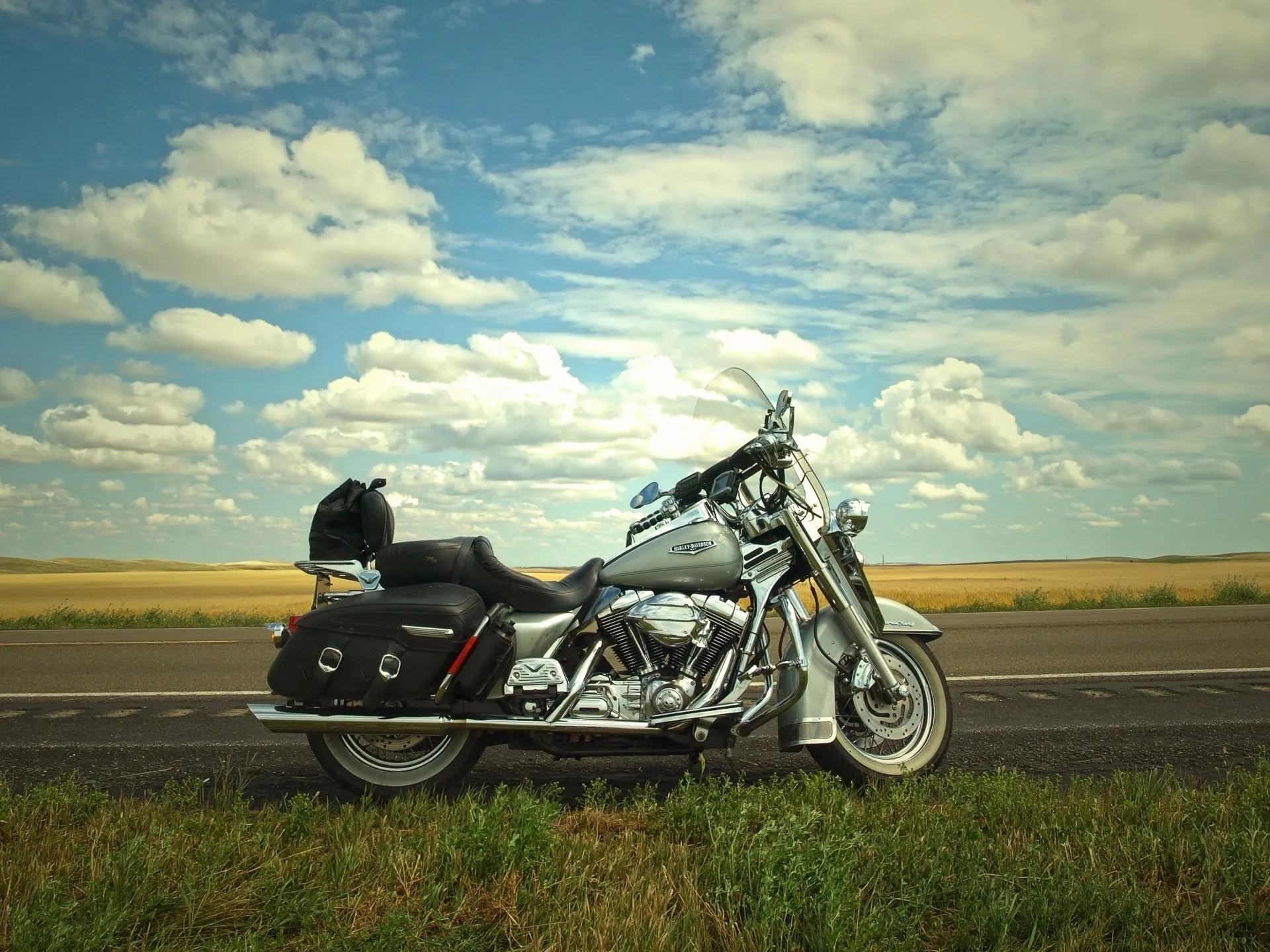 A silver motorcycle parked on the side of a rural road with a cloudy sky and expansive fields in the background.